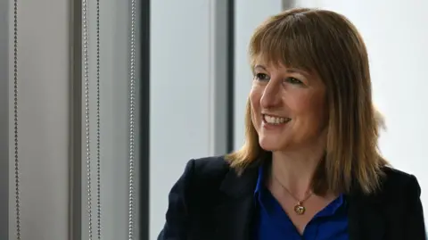 PA Media Rachel Reeves looks to the side smiling - she has shoulder-length brown hair and is wearing a black blazer with a blue shirt and a gold necklace