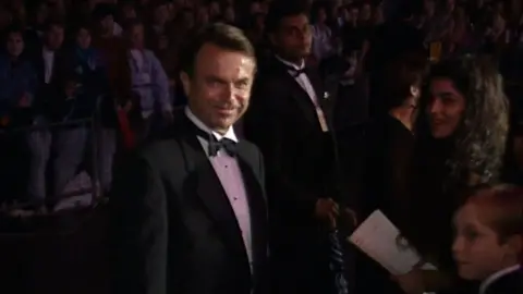 Sam Neill smiling and standing next to his young son during JP premiere in 1993