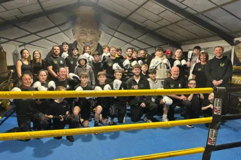 Cwmavon Hornets ABC The image shows a group of boxers in a boxing ring, posing for a photo while wearing new boxing gloves. 