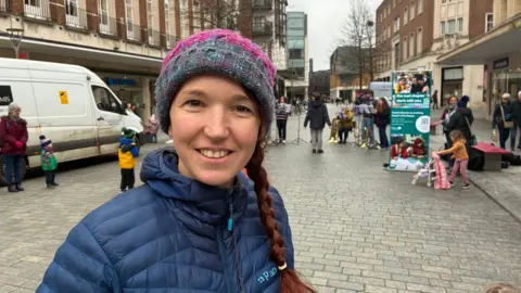 Hannah Rudd, wearing a grey and pink woolen hat with a single plait of hair falling over her shoulder. She is wearing a blue padded jacket and the brass band is playing behind her in Bedford Square.
