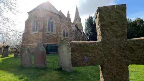 A large church with yellow cobbled brickwork standing in green grounds, with a graveyard full of monuments including grave stones and crosses.