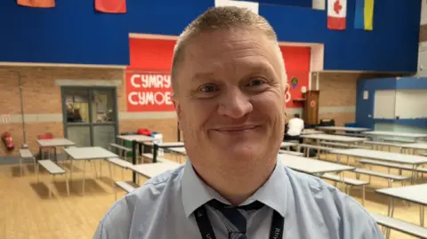 Gareth Jones smiling looking at the camera. He is stood in the school hall. He is wearing a light blue shirt and navy and white striped tie. He has short ginger hair.