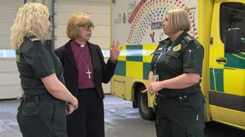 Archbishop of Canterbury Dame Sarah Mullally (centre) wearing dog collar and cross speaking to two Secamb female staff next to an ambulance