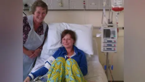 Jo Kelly A woman is sitting in a hospital bed with a blue long-sleeved top and yellow banana trousers. She has hospital equipment wrapped around her arm. Another woman is standing next to her above the bed in a plastic apron.