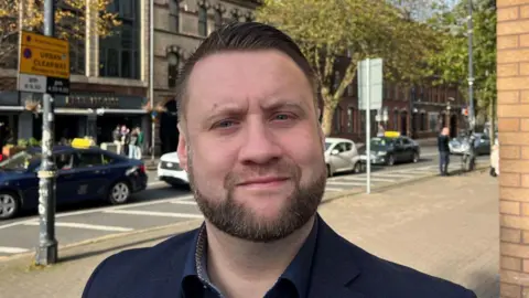 Mr Henderson wearing a navy shirt and navy suit jacket as he stands on a footpath. In the background there are five cars pictured as they queue and make their way down a street in the city.