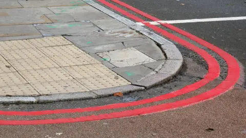 Double red line road markings on the corner of a road. Double red line markings means no parking at any time. The pavement slabs are cracked and the road surface has been patched up.