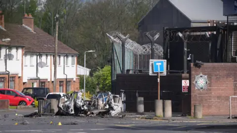 Niall Carson/PA Wire The wreckage of a car bomb outside a police station, with a row of terraced houses and trees in the background.  The car has been blown apart and lies on the road beside a set of pavement bollards. The station is protected by a large metal fence, topped with barbed wire, and a red brick wall.  