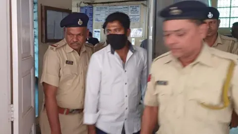 Mukesh Kumar A man, wearing a white. open-neck shirt and a black face mask, is escorted by several Indian policemen wearing beige uniforms and dark-coloured berets.