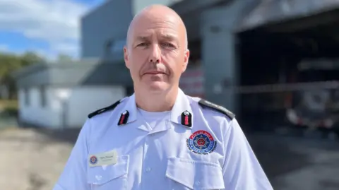 Mark Christian is wearing a white fire service shirt, and he has no hair. He is standing in front of the burned fire station on a sunny day.