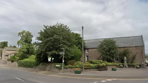 Google View of a small public space on a street corner, with a low wall and a hedge behind two seating benches.