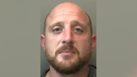 A mug shot of a bald man with a light-brown beard looking into the camera.