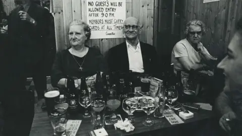 Paul Trevor A black and white image of an elderly man and woman sitting at a table in a pub - the table in front of them has many glasses on it. 