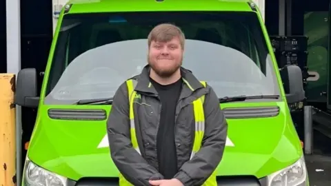 ASDA Joe, who has brown hair and a beard, sits on the bonnet of a green Asda delivery van with his almost crossed smiling for a photograph. He is wearing a dark jacket and high-vis vest.