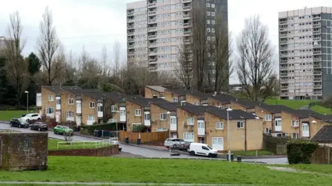 Two grey tower blocks stand behind two rows of small, beige maisonettes. They stand on a slight hill with a road and then grass in front and behind them by the tower blocks.