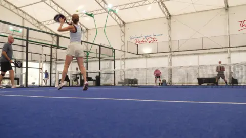 A team of two men and two women play padel. The indoor court looks much like a tennis court. A woman is leaping up to hit back a ball.