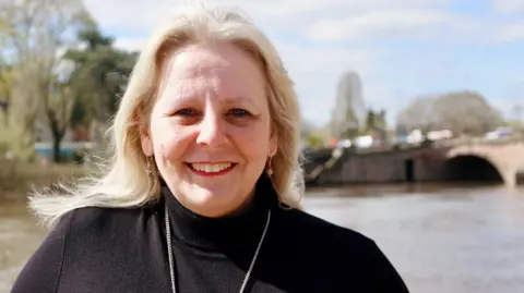 LDRS Elena Round wears a black turtle necked jumper with a silver chain. She has long wavy blonde hair.