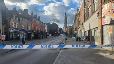 BBC Blue and white "Police" tape is across both ends of the road. A police officer is stood just behind the tape to the right of the picture. Two other officers are kneeling on the floor in the background.