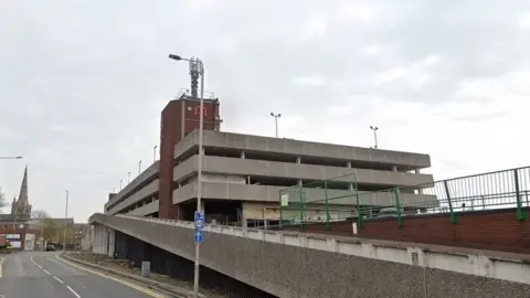 A brutalist-style concrete multi-storey car park viewed from the roadside entrance ramp