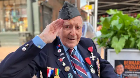 An older man, in a military cap, with numerous badges and medals on his blazer, salutes at the camera. There is a poster behind him featuring his name and a picture of him