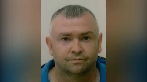 PSNI Damien Heagney, wearing a blue collared shirt in front of a grey background. 