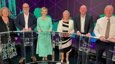 Three men and three women stand behind glass podiums in a TV studio in front of a green and purple backdrop. Sally Horscroft, on the left, wears a floral dress and dark jacket. Jason Stockwood, next left, wears a dark jacket and open-neck white shirt. Dame Andrea Jenkyns wears a light green dress. Marianne Overton wears a black and white dress and cream jacket. Rob Waltham wears a dark suite and open-neck white shirt. Trevor Young wears a white shirt and striped tie.