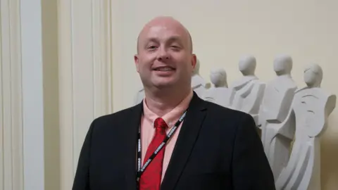 LDRS Councillor Paul Stuart. He is smiling, has no hair, and is wearing a black jacket, pink shirt, and red tie.