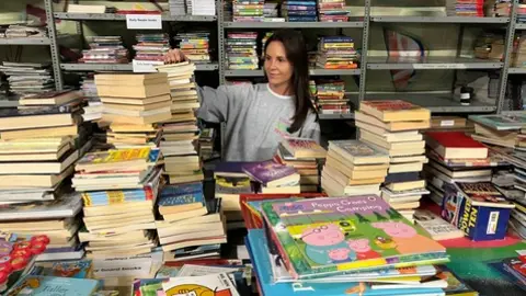 The Children's Book Project A woman with long brown hair and a brown jumper stands in a room surrounded by books