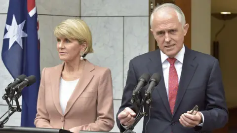 Getty Images A woman, with short blond hair and in a light suit, and a man in a dark suit, both standing at a podium with a partial Australian flag in background.