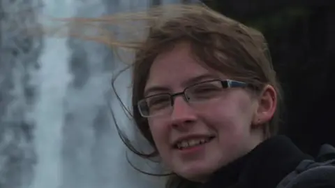 Headshot picture of a woman wearing glasses and brown hair who is smiling. Her hair is tied back but is windswept. She is stood outside and is wearing a black coat.