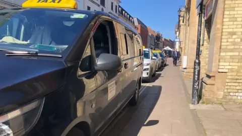 Vikki Irwin/BBC Black and white taxis are lined up on the taxi rank in Bury St Edmunds. It's a sunny day with blue skies.