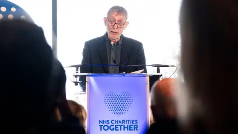 PA Media Michael Rosen, a man wearing a black suit and grey shirt, is stood at a blue lectern which has the words NHS Charities Together written on it along with a heart symbol.