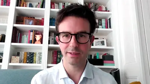 A man, Aron Gelbard, is seated on a sofa in front of a bookshelf filled with various books and decorative items. He is wearing a light-colored shirt, and a yellow pillow is visible to his right. The background is in focus, showcasing the bookshelf’s contents clearly.