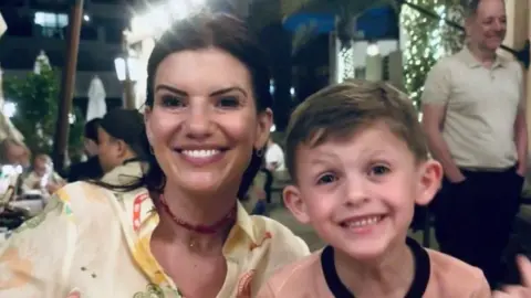 British Heart Foundation Lucie and Archie sitting next to each other at a dinner table outdoors. They are both smiling. Lucie is wearing a pale yellow shirt and has black hair. Archie is wearing a pale pink top with a black collar.