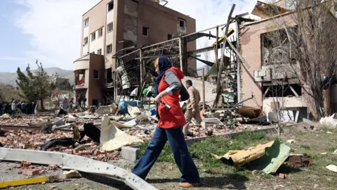 EPA A woman in a red top carrying a microphone walks through debris in front of a damaged building at the Shahid Beheshti University, in Tehran.