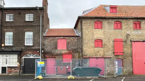 Clare Worden/BBC A row of brick buildings. The doors and windows of the Sommerfeld and Thomas warehouse are painted red. The smaller building, of about two storeys, is the one which is being renovated. It has a red door and shutters.