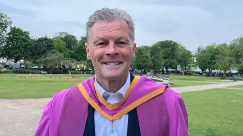 Cheltenham Borough Council Steve, a middle-aged man, smiling to camera in a purple graduation gown over a light blue shirt and dark jacket. There is a landscaped public garden behind him and a row of trees in the distance.