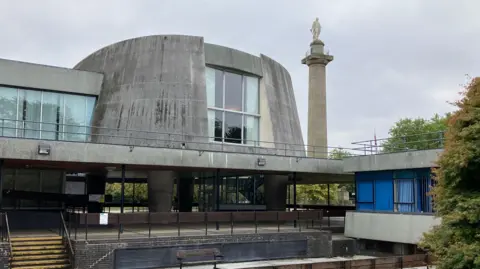Steps leading up to an oval council chamber on large concrete stilts. A tall column with the statue of a man can be seen in the background.