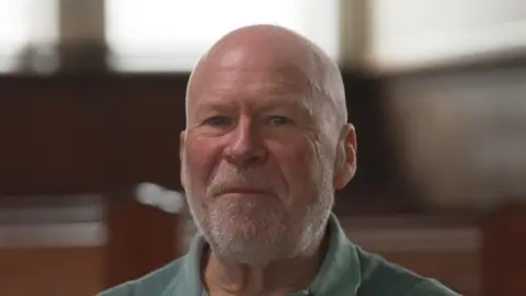 A bald man with a short grey beard is looking at the camera. He is wearing a green polo-neck tshirt. Behind him is a large, out of focus room, which appears to show church pews. 
