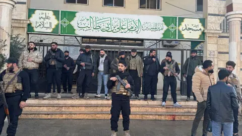 Omar HAJ KADOUR / AFP via Getty Images about 17 men with guns stand outside a building with a big green sign.
