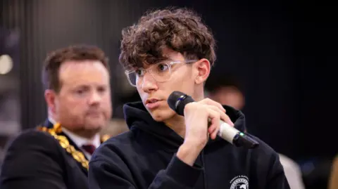 ISCRE A young man looks to one side as he holds a microphone. An older man wearing a ceremonial chain around his neck looks on in the background. The young man is wearing a black hooded sweatshirt. 