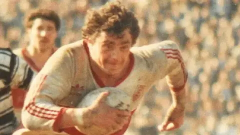 An archive image of Phil Lowe, wearing a red and white Adidas kit. He is carrying a rugby ball with a determined expression on his face. 