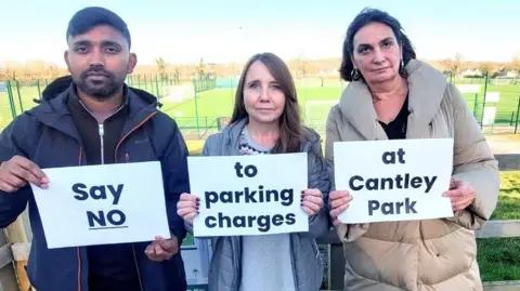 Three Labour councillors standing in Cantley Park car park. Small football pitches are visible behind them. They each hold a banner, which when read in order are 'Say NO' 'to parking charges' 'at Cantley Park'.