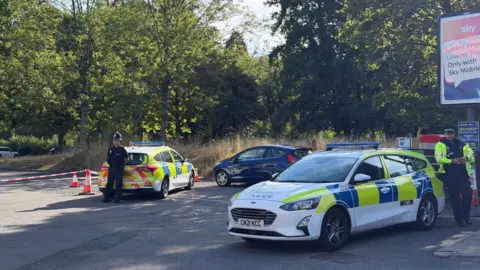 A police cordon with two police cars and a police officer.