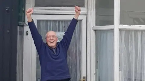 Dorothy Lambert Charlie Lambert celebrating outside Norman Nicholson's house after the property was bought in February. Mr Lambert is smiling and his hands are up in the air. he is wearing a navy blue sweatshirt and has glasses. The house has large front windows with white panes. 