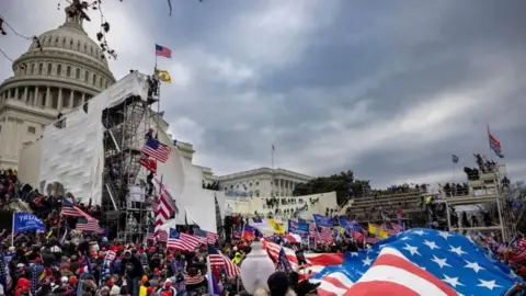 Crowd scene outside the US Capitol building on 6 January 2021. A mass of people wave US flags and Trump banners under a darkened sky