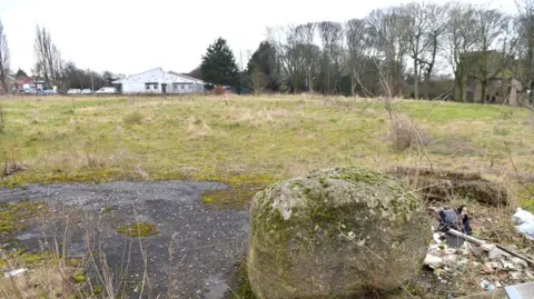 LDRS A boulder in the foreground and green scrubland with a white building (the swimming pool) in the distance to the left and part of Bolling Hall Museum to the right. 