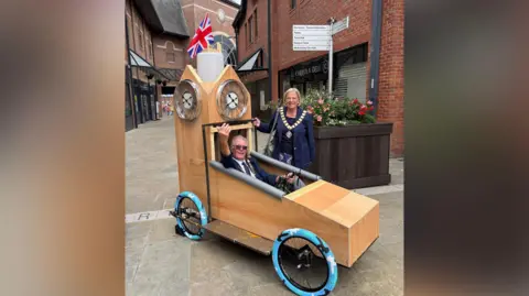 Barrow BID Mr Chatfield sits in the cart with one hand on the wheel and the other raised. Another woman wearing a ceremonial gold necklance stands next to them, smiling broadly. The card has tall wooden panelling that resembles a clock tower. A union jack flag is placed on top.