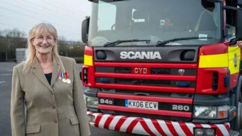 Northamptonshire Fire and Rescue Service A woman in a grey suit jacket with three medals stands next to a red fire truck.