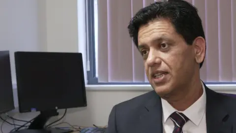 Dr Zahid Chauhan has short dark hair, a grey jacket, white shirt and black tie with white and red stripes.