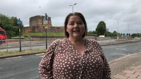 BBC Julie Minns outside the BBC Radio Cumbria office in Carlisle wearing a spotted brown blouse and smiling. There is a road and the castle in the background. 